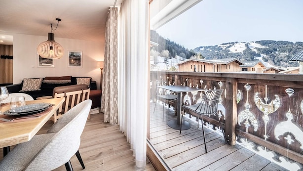 Un balcón con barandilla de madera, una mesa de comedor con sillas y vistas a casas y montañas nevadas.
