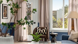 Quarto com vista para árvores através de uma janela, uma poltrona marrom com almofada xadrez, um sofá azul, uma mesa com abajur e um quadro na parede.