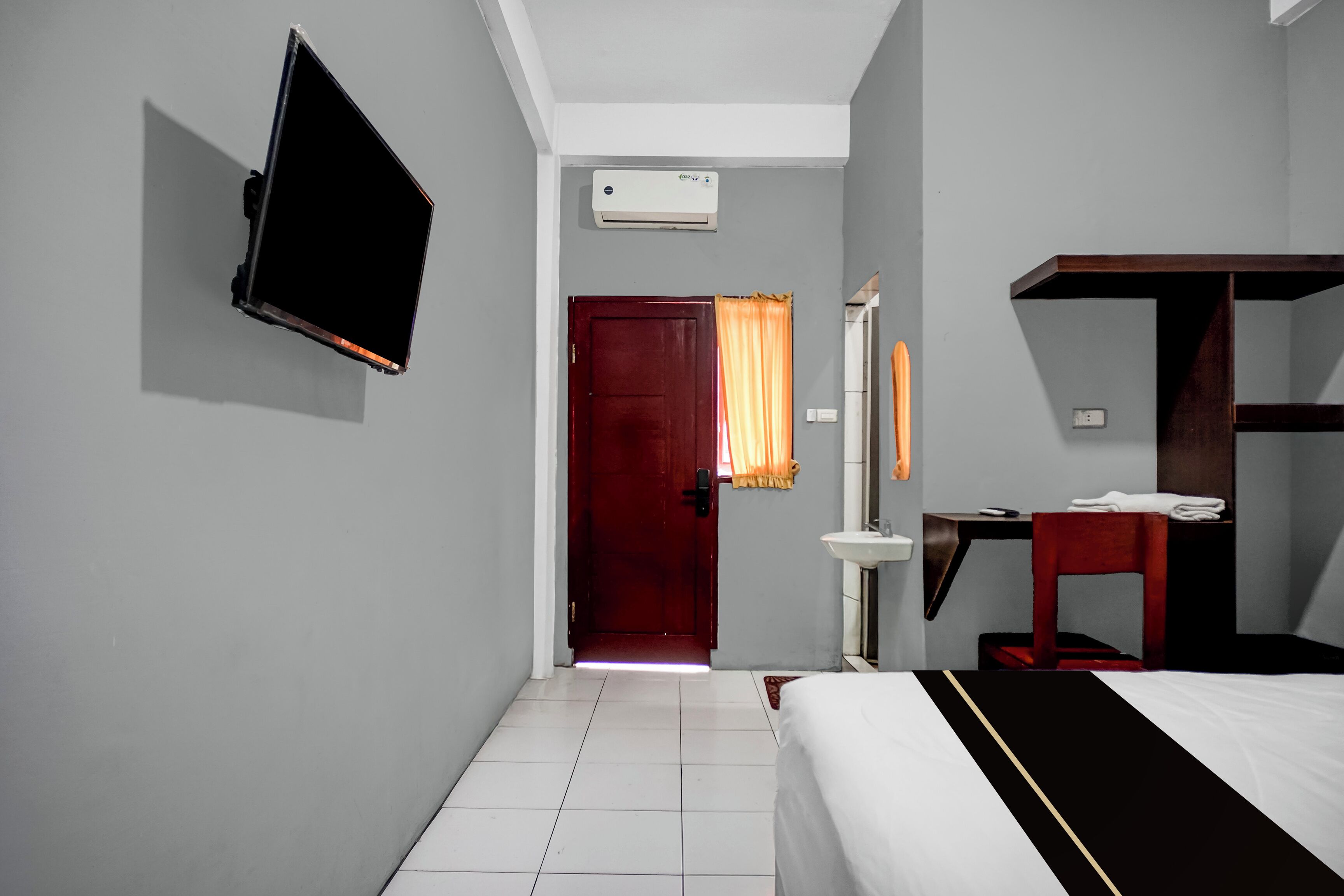 A hotel room with a bed, a TV mounted on the wall, a desk with a chair, a mirror, and an air conditioning unit.