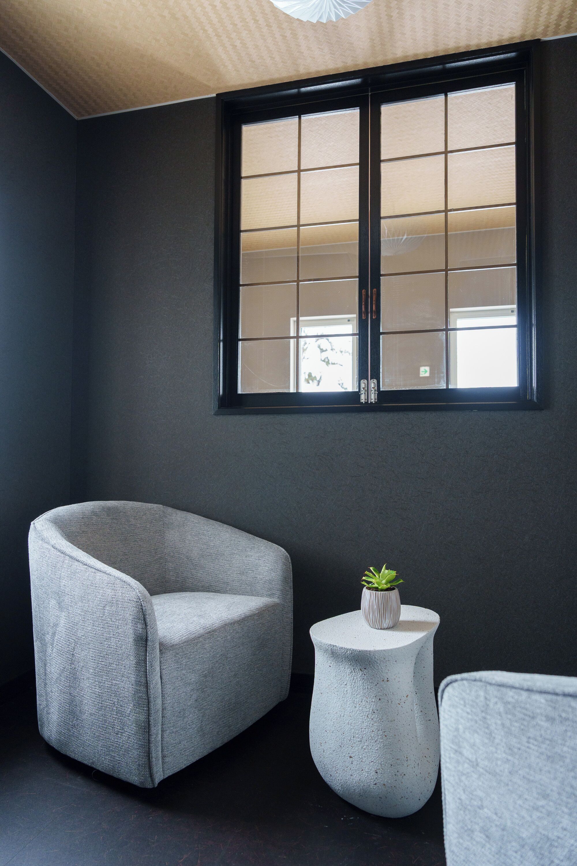 Ein Zimmer mit einer dunkelgrauen Wand, einem großen Fenster mit Gittermuster, einem grauen Sessel, einem kleinen runden Tisch mit einer Pflanze und einem weiteren grauen Stuhl.