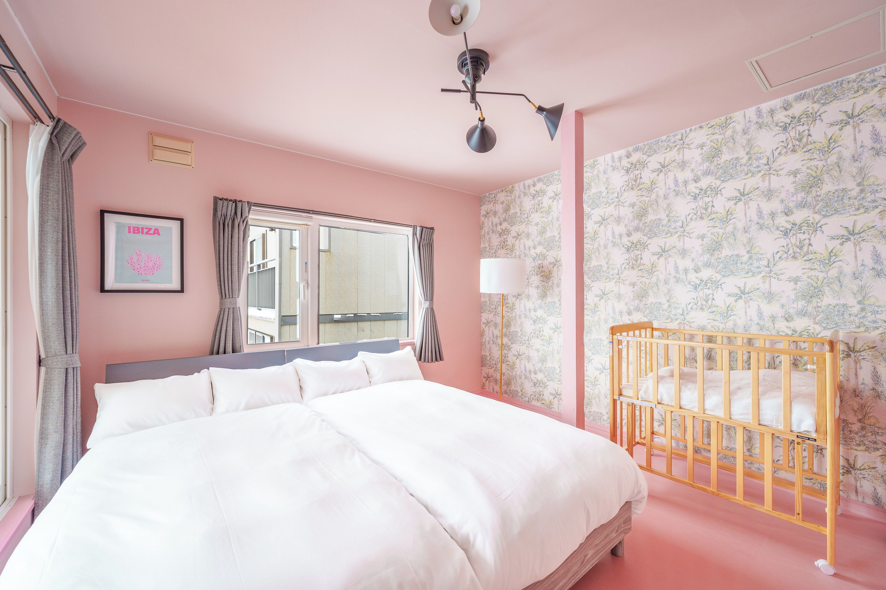A pink-themed hotel room with a large bed, a crib, a window with curtains, and a wall-mounted picture frame.