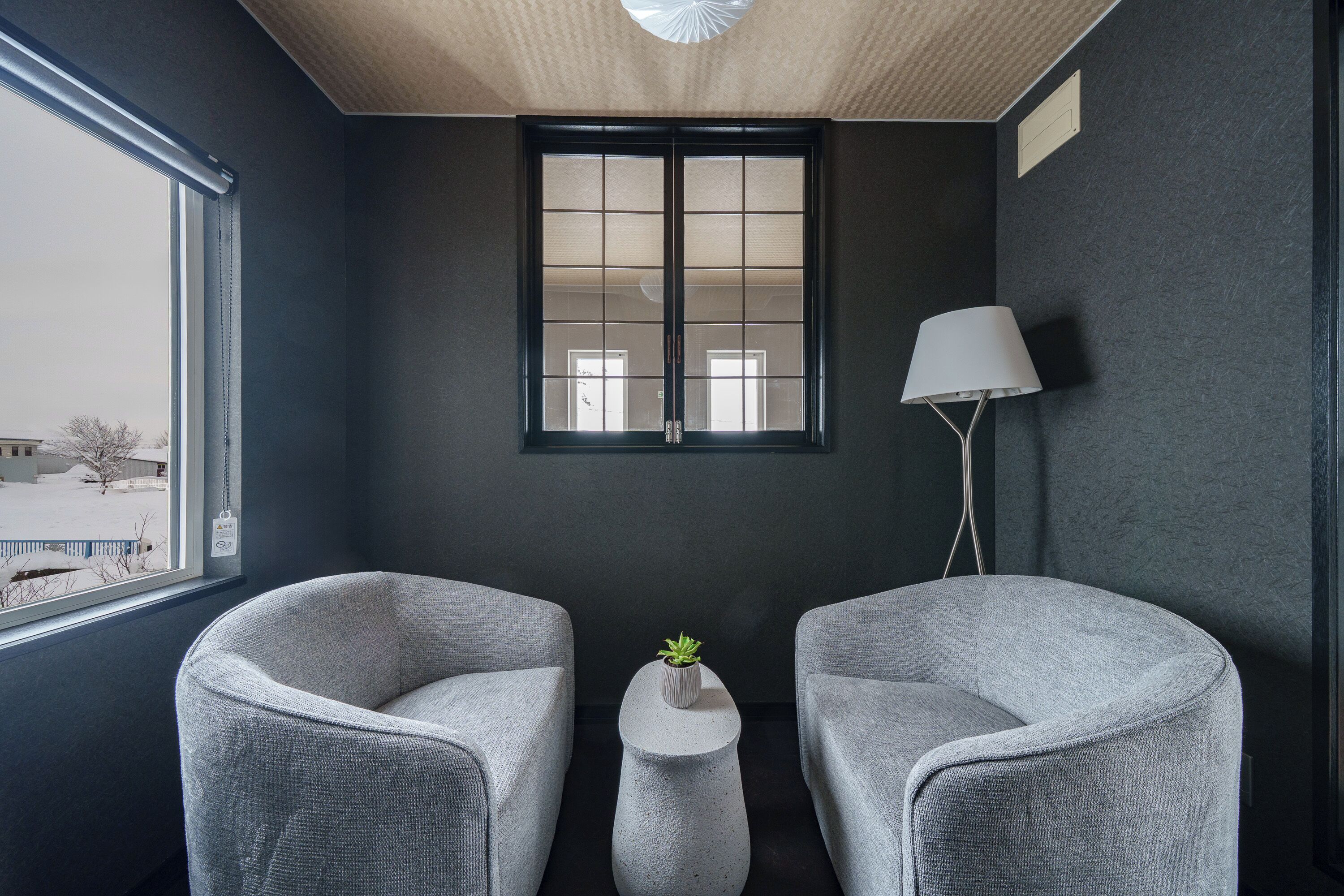 A room with two grey armchairs, a small white table with a plant, a floor lamp, and a window with a view of a snowy landscape.
