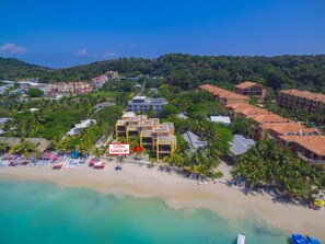 Vista aérea de un complejo turístico costero con varias construcciones, una playa de arena y abundante vegetación.