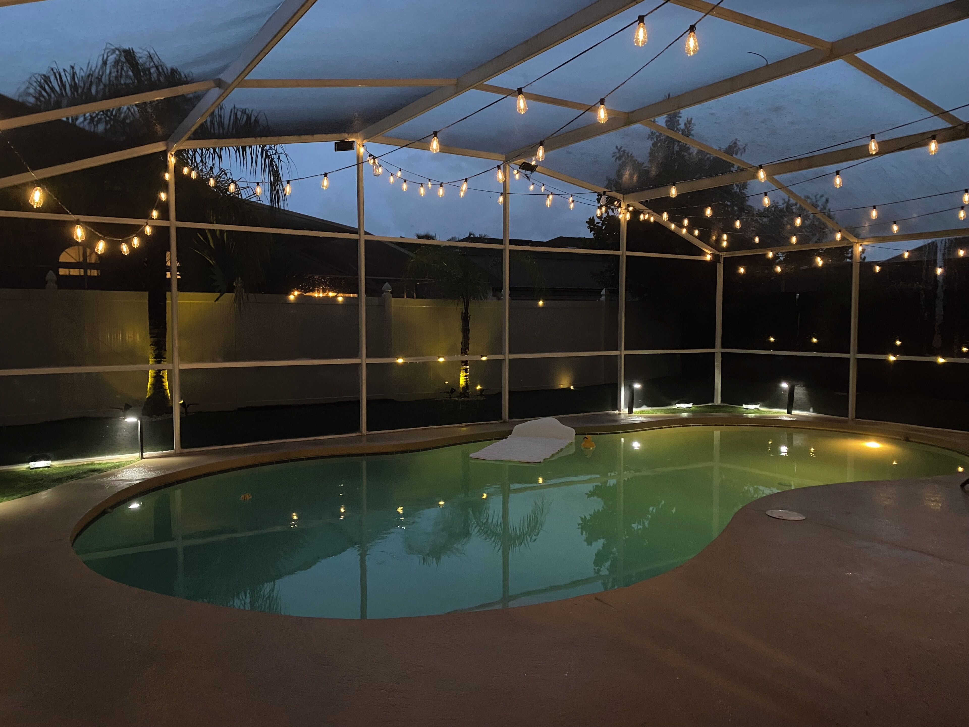 A swimming pool with string lights, surrounded by a covered area with a view of the sky.