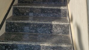 Una escalera con escalones de mármol oscuro y barandilla de madera, que asciende por un estrecho pasillo con paredes lisas.