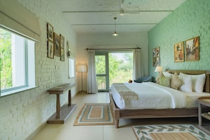 A bedroom with a bed, a bedside table, a lamp, a window, and a view of greenery outside. - Kadamb Bagh (Rajgarh)