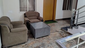 Une pièce comprenant un canapé, un fauteuil et une petite table. On y trouve une porte en bois et une fenêtre avec des rideaux. Un escalier est visible au fond de la pièce.