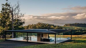 A modern glass house with a flat roof, situated in a grassy field with a view of distant hills and trees. - Triptych - The Glass House (Tasman)