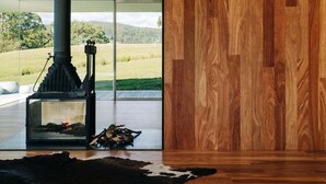 A modern interior with a wood-burning stove, glass doors, and a view of a grassy landscape. - Triptych - The Glass House (Tasman)