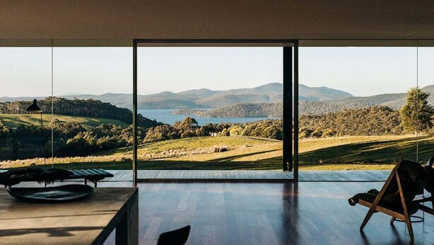 A room with a large glass door leading to a scenic outdoor view of hills and a body of water. - Triptych - The Glass House (Tasman)