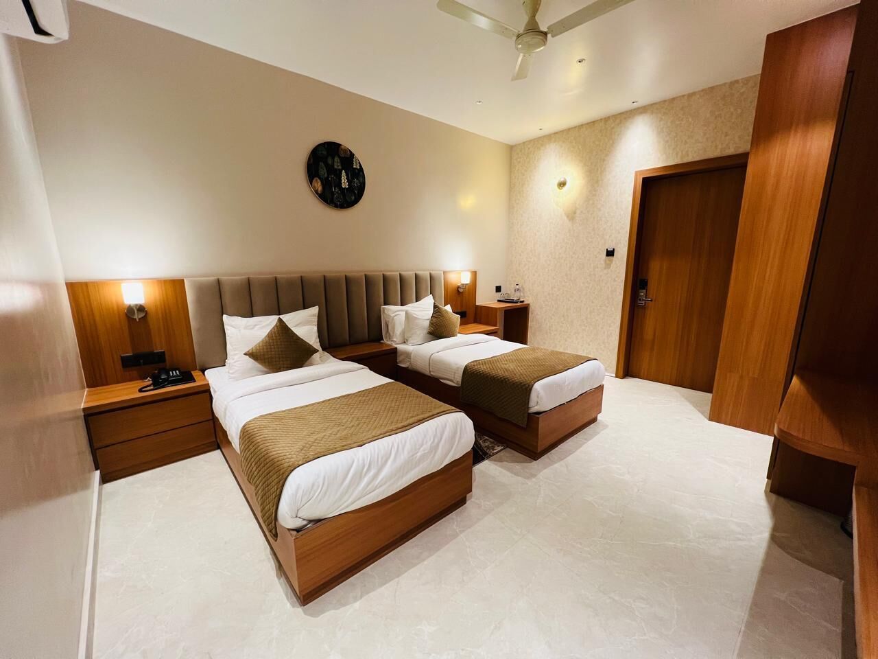 A hotel room with two beds, a wooden headboard, a nightstand with a lamp, a clock on the wall, and a door.