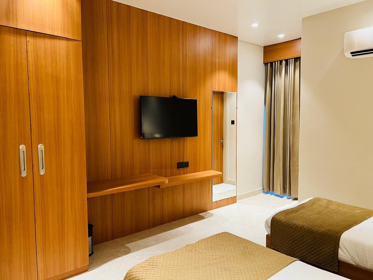 A hotel room with a wooden wardrobe, a flat-screen TV mounted on the wall, a bed with a brown blanket, and a window with curtains.
