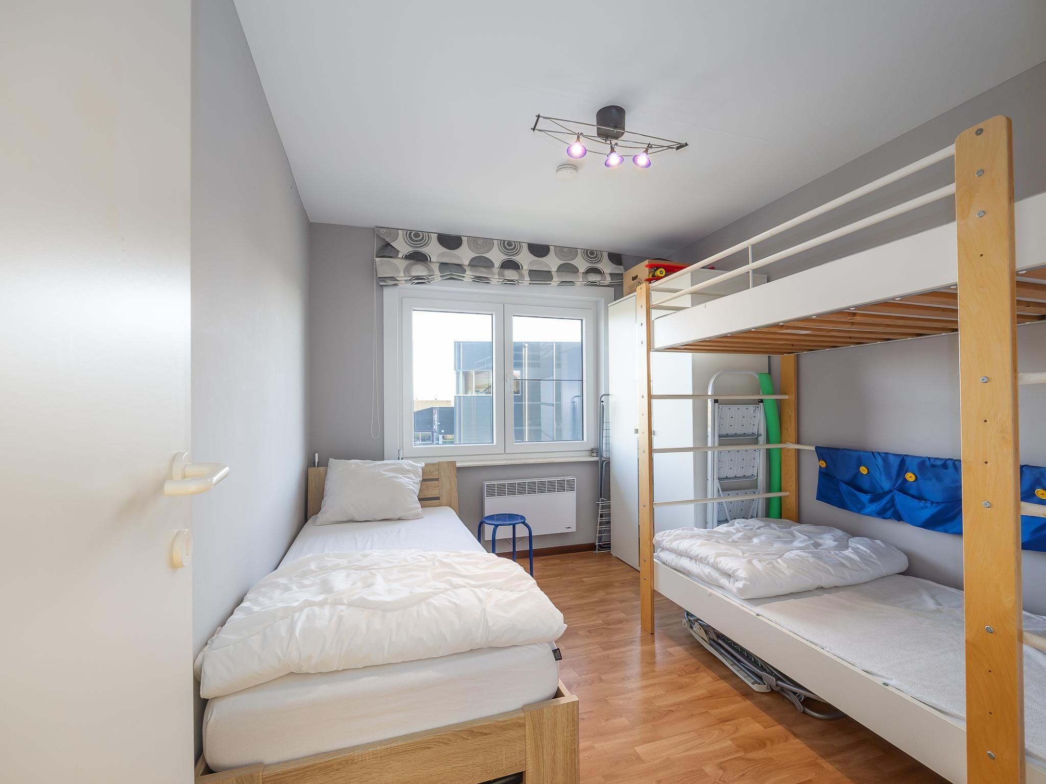 Een studentenkamer met stapelbedden, een raam, een radiator en een plafondlamp.