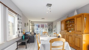 A modern kitchen with a dining area, featuring a large wooden cabinet, a dining table with chairs, and a view through the glass door to the outside. - Bright apartment with 2 bedrooms and garage (Middelkerke)