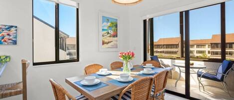 A dining area with a table set for four, surrounded by wicker chairs with blue cushions, a hanging wicker chandelier, and a view of the outside through large windows.