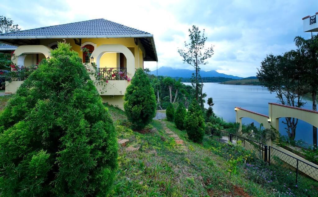 A house with a balcony overlooking a lake, surrounded by greenery and mountains in the background.