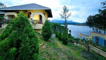 A house with a balcony overlooking a lake, surrounded by greenery and mountains in the background.
