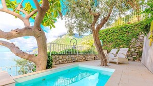 Una piscina exterior con sillones de descanso, rodeada de árboles y un muro de piedra, con vistas a un paisaje montañoso.