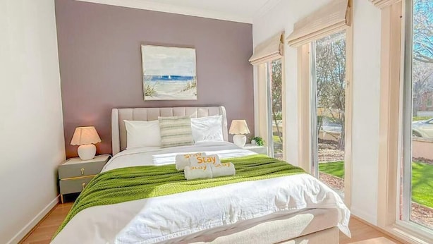 A bedroom with a bed, nightstands, a painting, and a view through large windows. - StayAU Family Haven Pet-Friendly (Caroline Springs)