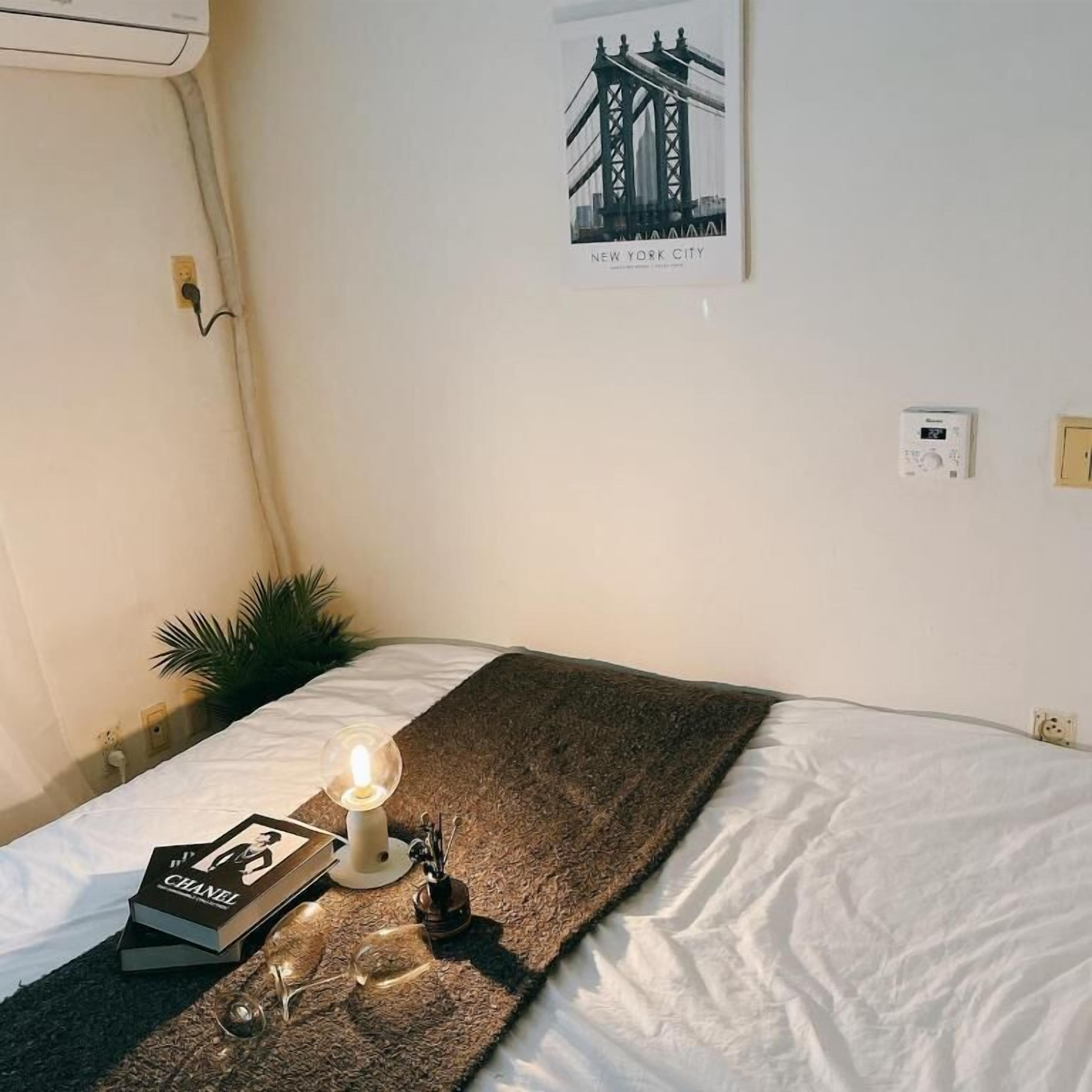 A neatly made bed with a white comforter and a brown throw blanket. A book titled "CHANEL" and a pair of glasses are placed on the bed. A lit lamp is on the bedside table. A poster of the Brooklyn Bridge is on the wall.