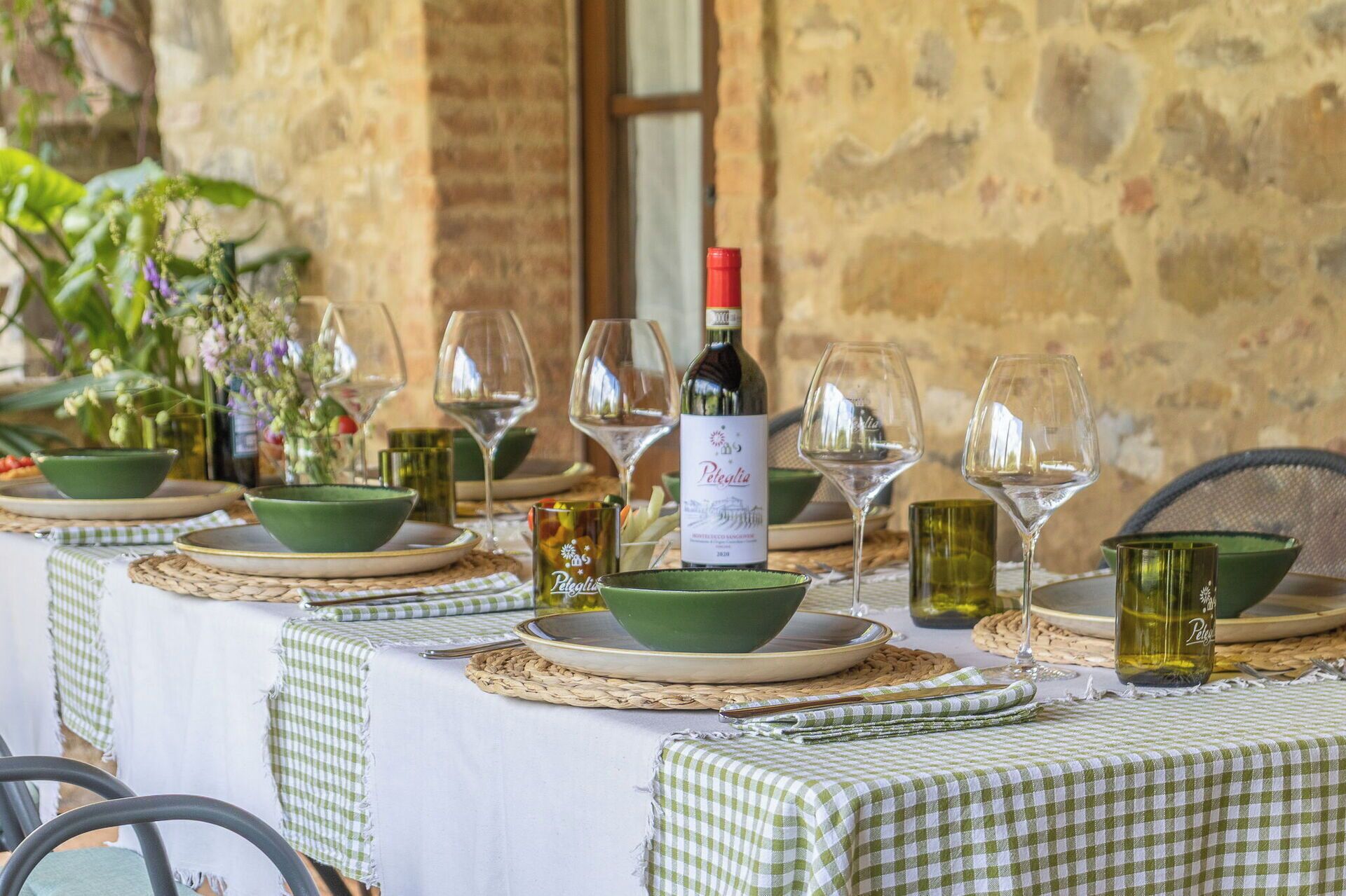 Une table dressée avec des verres à vin, des bols et une bouteille de vin, recouverte d’une nappe à carreaux et agrémentée de sets de table tissés.