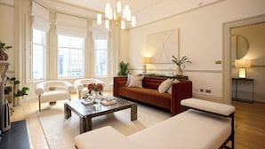 A spacious living room with a large window, a modern chandelier, a red sofa, and a white armchair. - Kensington Five-Bedroom Townhouse (London)