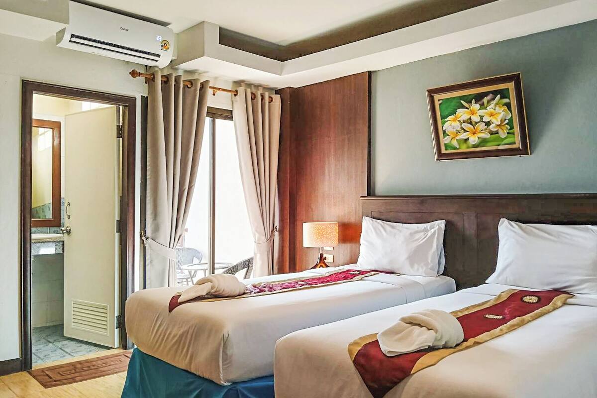 Une chambre d’hôtel avec deux lits, un tableau représentant des fleurs accroché au mur, une fenêtre avec des rideaux et un appareil de climatisation.
