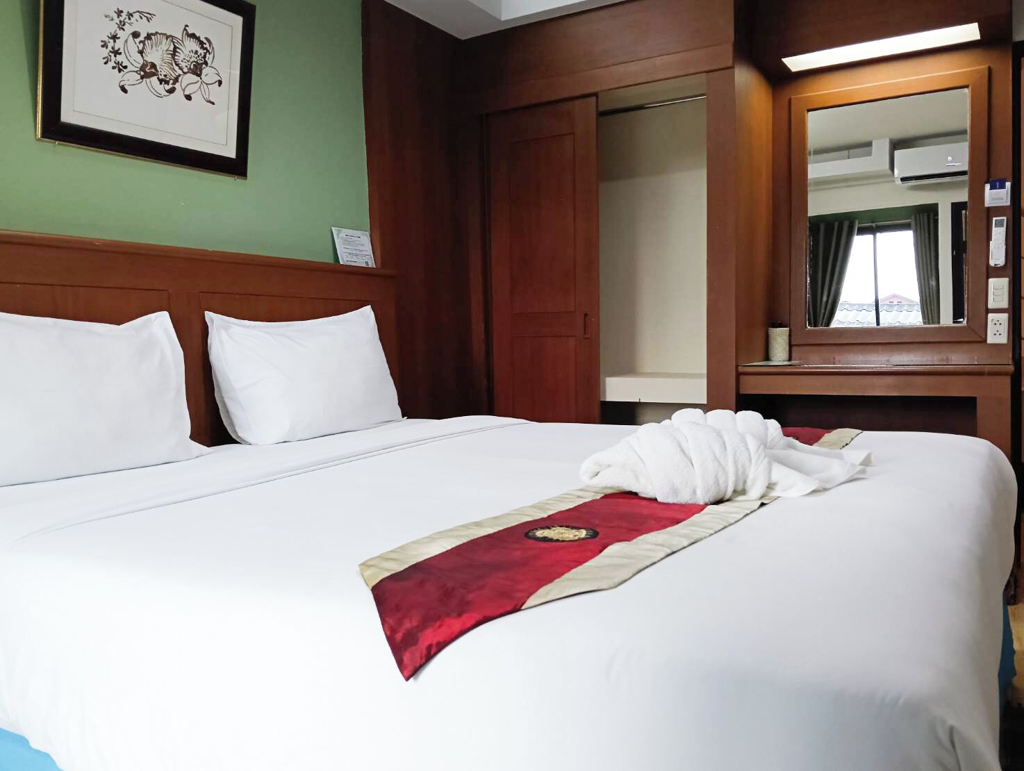 A hotel room with a large bed, two white pillows, a red and beige bed runner, a framed picture on the wall, and a mirror above the bed.
