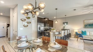 Una cocina moderna con electrodomésticos de acero inoxidable, un comedor con mesas de vidrio y sillas de madera, y una sala de estar con un sofá y un cuadro.
