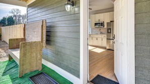 Une façade en bois agrémentée d’une lanterne en métal, un paillasson vert à l’entrée et, à l’intérieur, une cuisine avec des meubles blancs et un réfrigérateur en acier inoxydable.