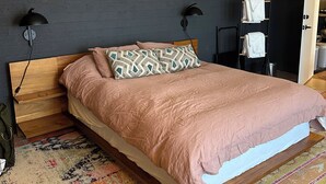 Une chambre avec un lit en bois, des coussins à motifs, un mur foncé, un ventilateur de plafond et un porte-serviettes suspendu.