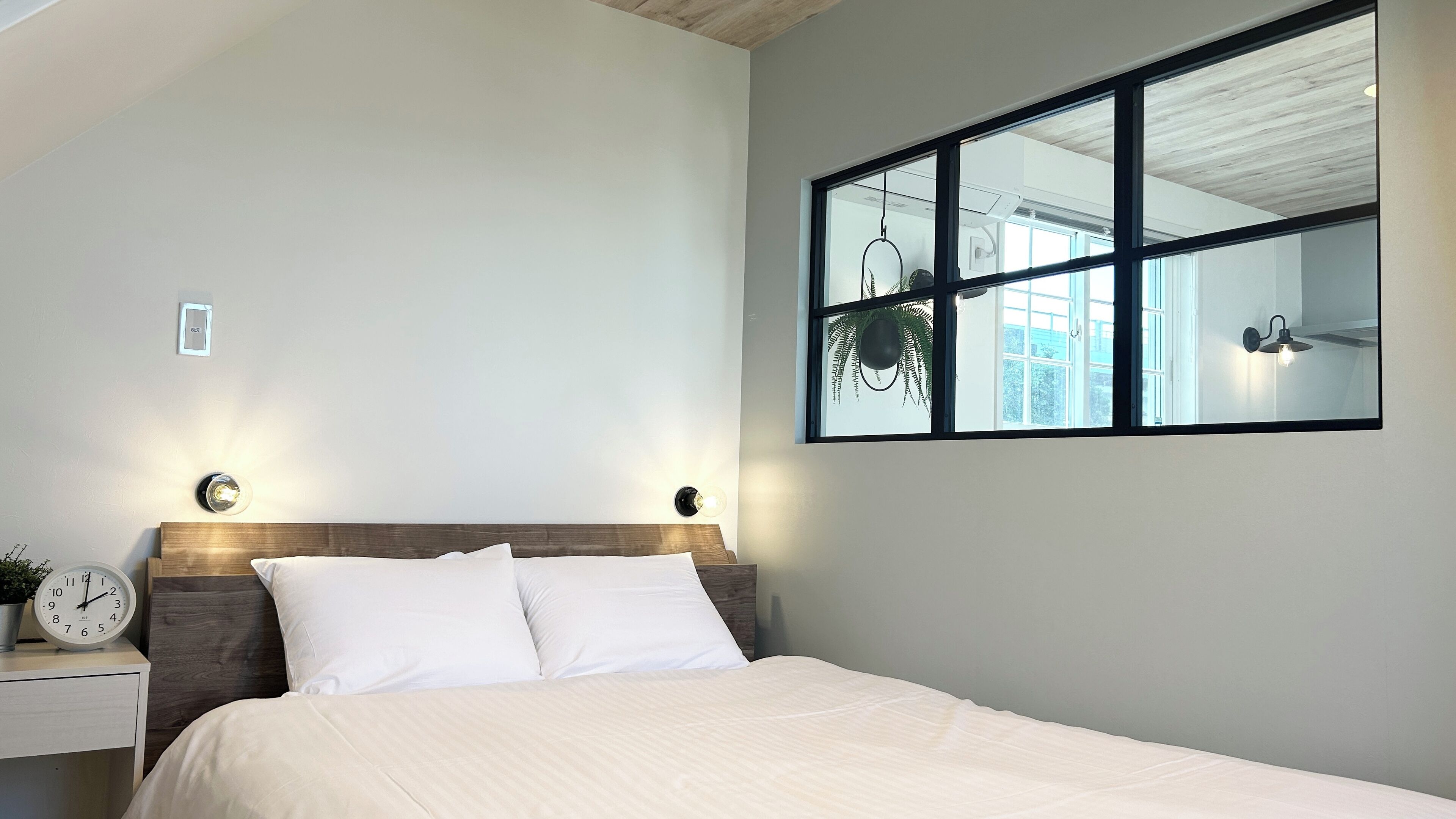 Un dormitorio con cama, mesita de noche con reloj y espejo en la pared.