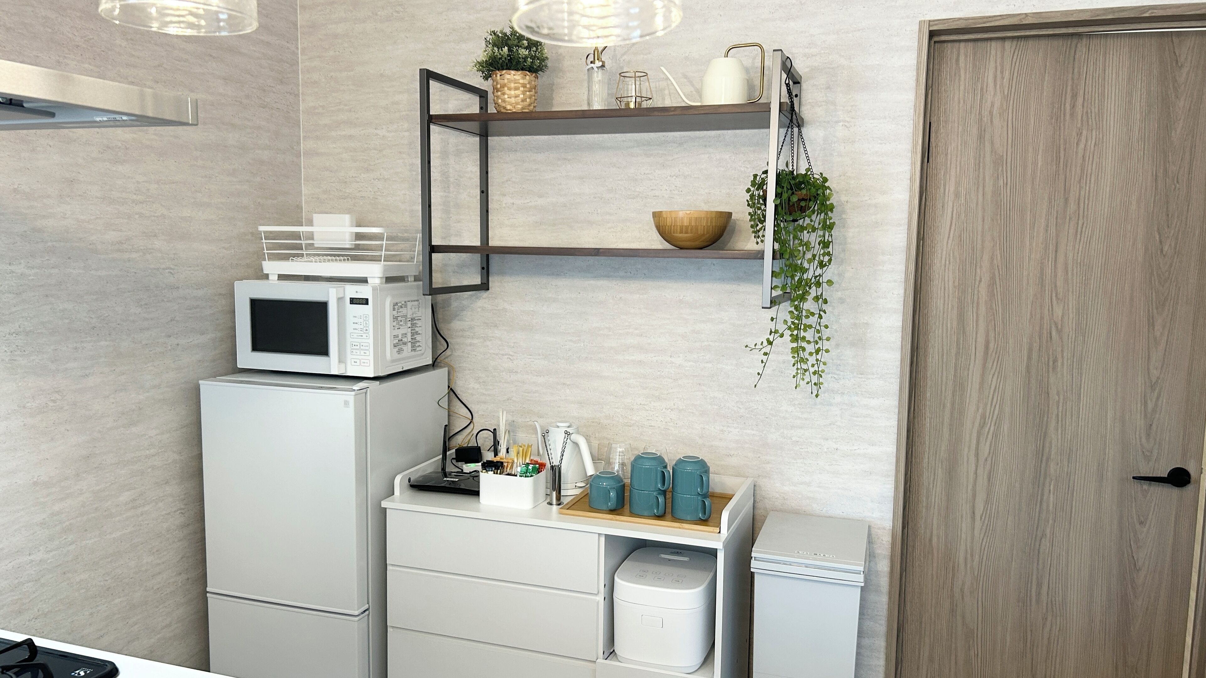 A compact kitchen with a microwave, a small refrigerator, and a coffee machine. There are shelves with plants and a tray of cups.