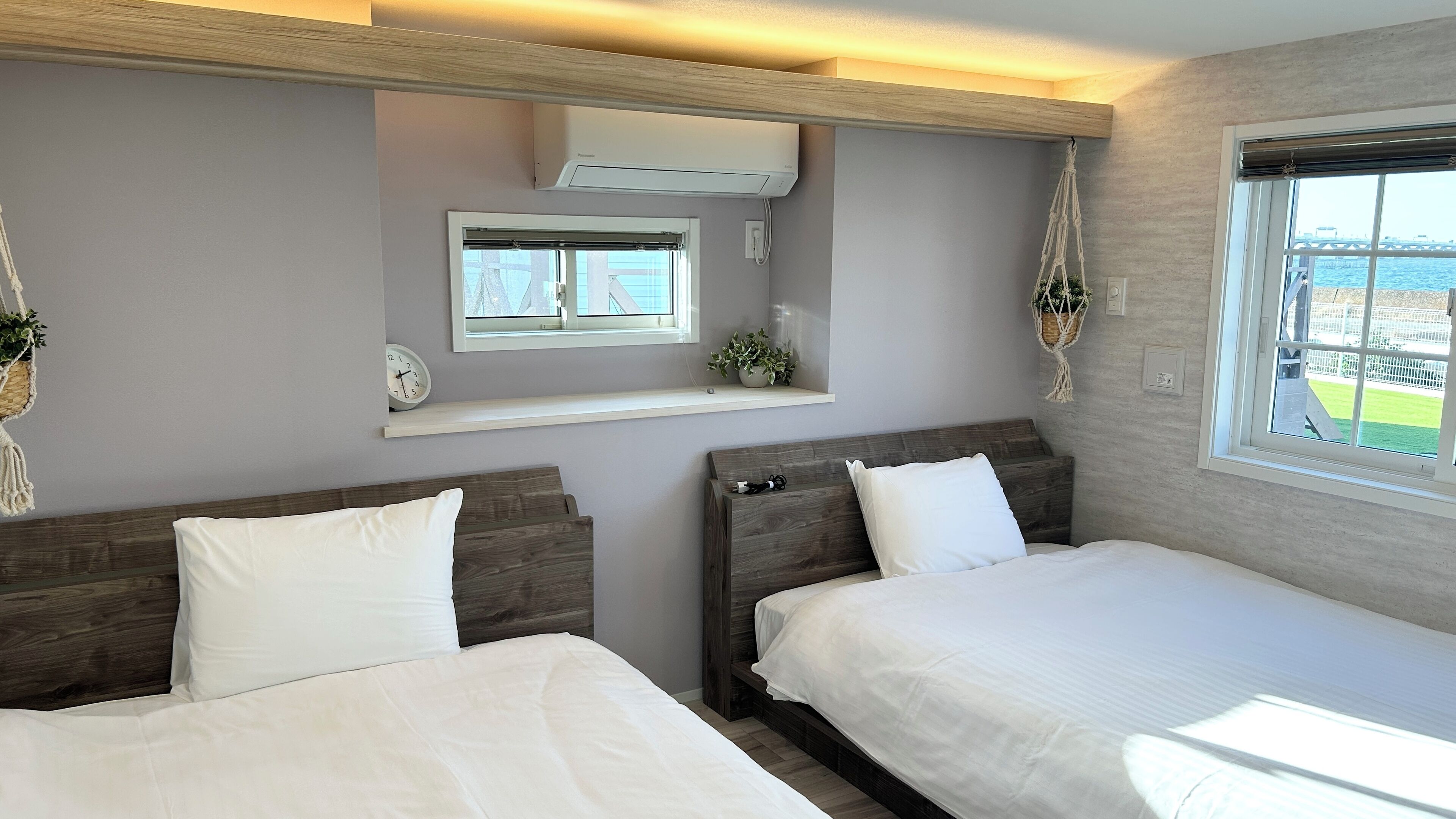 A hotel room with two beds, a window with a view, an air conditioning unit, a shelf with a plant, and a macrame hanging.