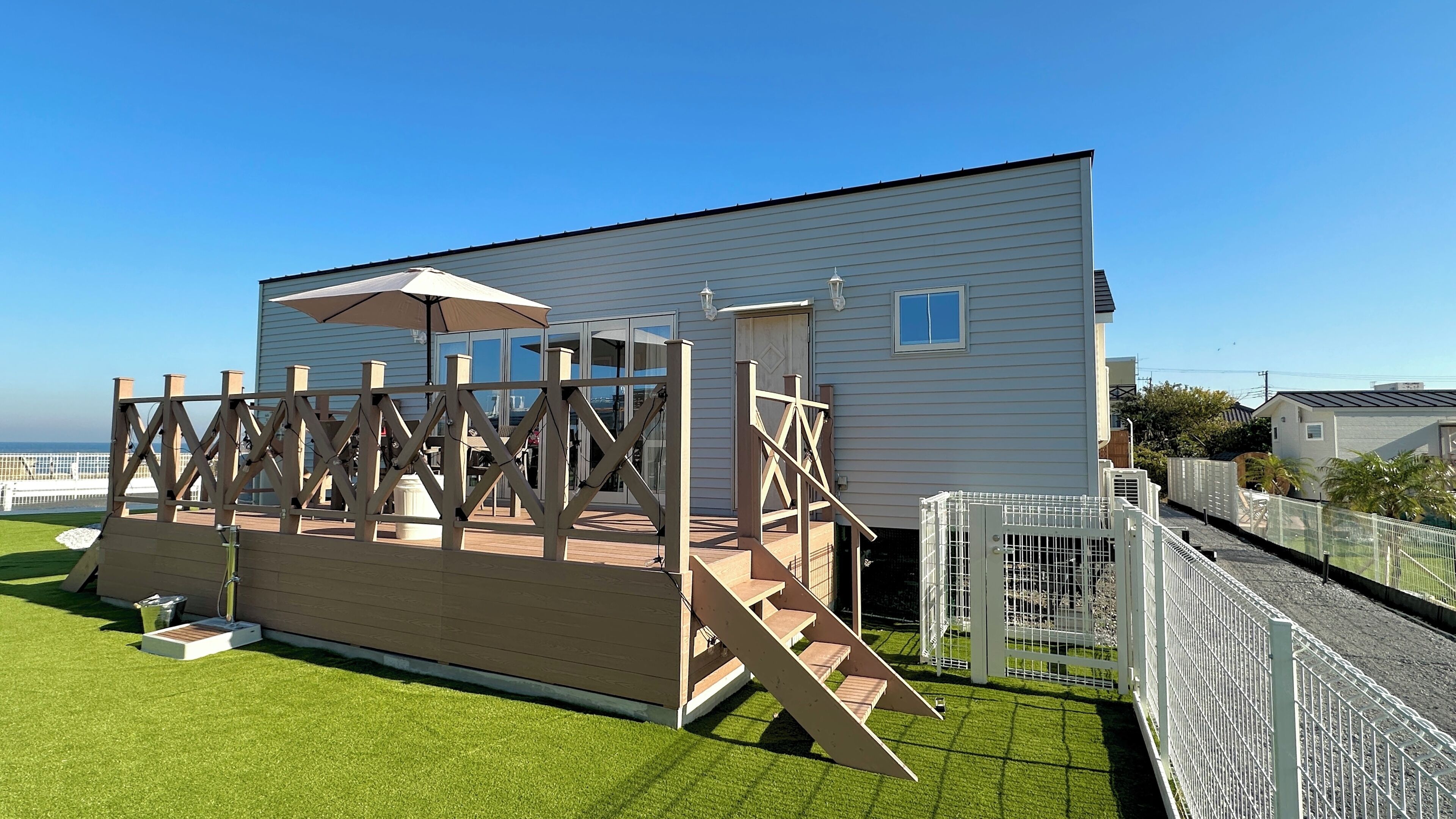 Eine Holzterrasse mit einer Pergola und einem überdachten Sitzbereich, angrenzend an ein weißes Gebäude mit einem großen Fenster und einer Tür.