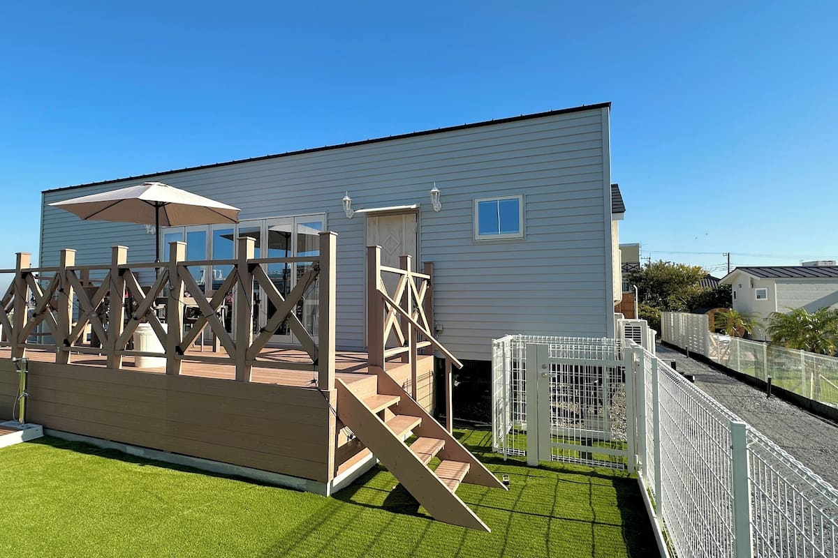 A wooden deck with a pergola and a covered seating area, adjacent to a white building with a large window and a door.