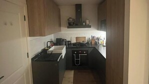 Cucina stretta con isola centrale, piano cottura e lavello. Sopra il piano cottura c'è un forno a microonde e uno scaffale con utensili da cucina. La cucina ha mobili in legno e uno schienale bianco.