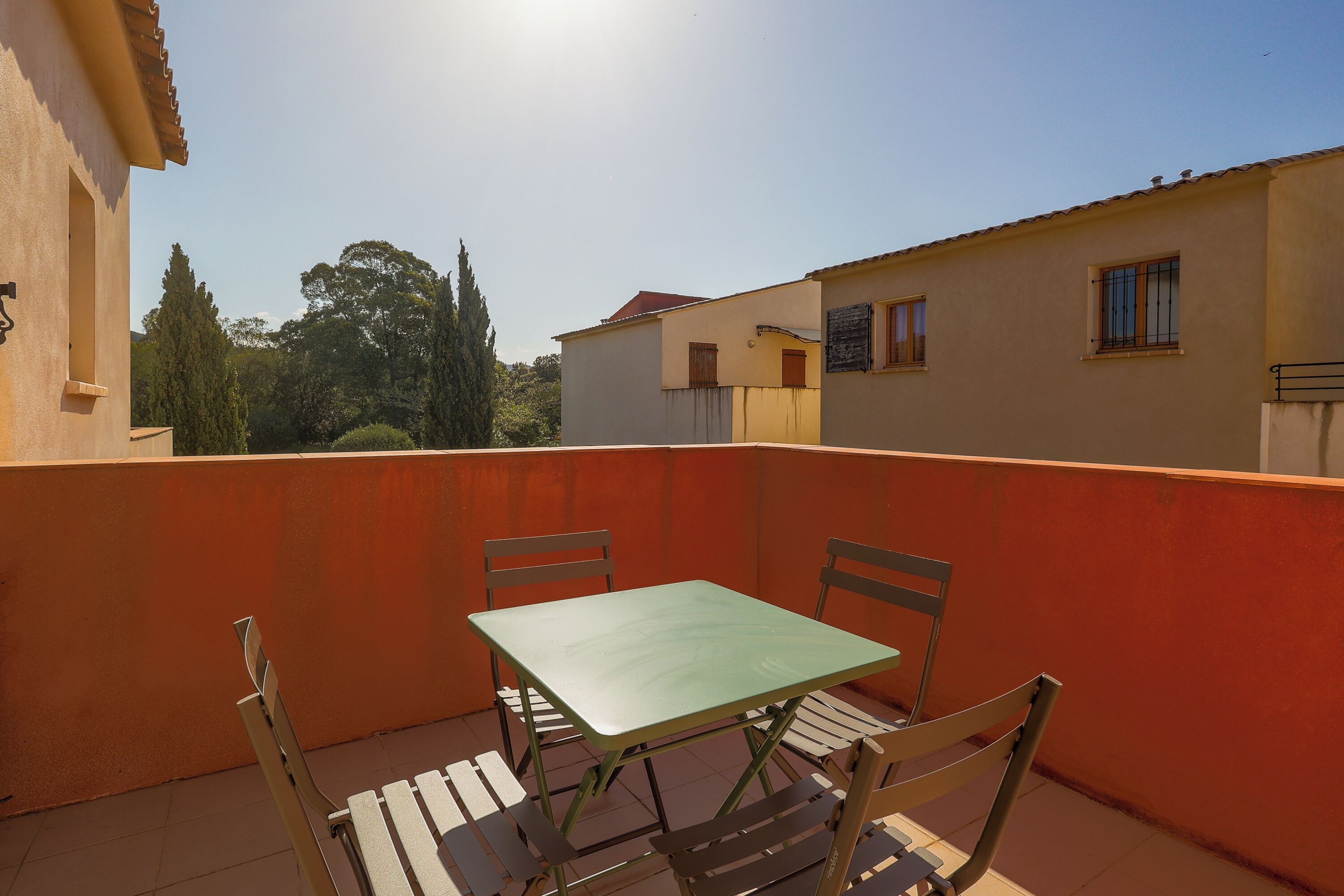 Een balkon met een tafel en stoelen, een betegelde vloer en uitzicht op omliggende gebouwen en bomen.