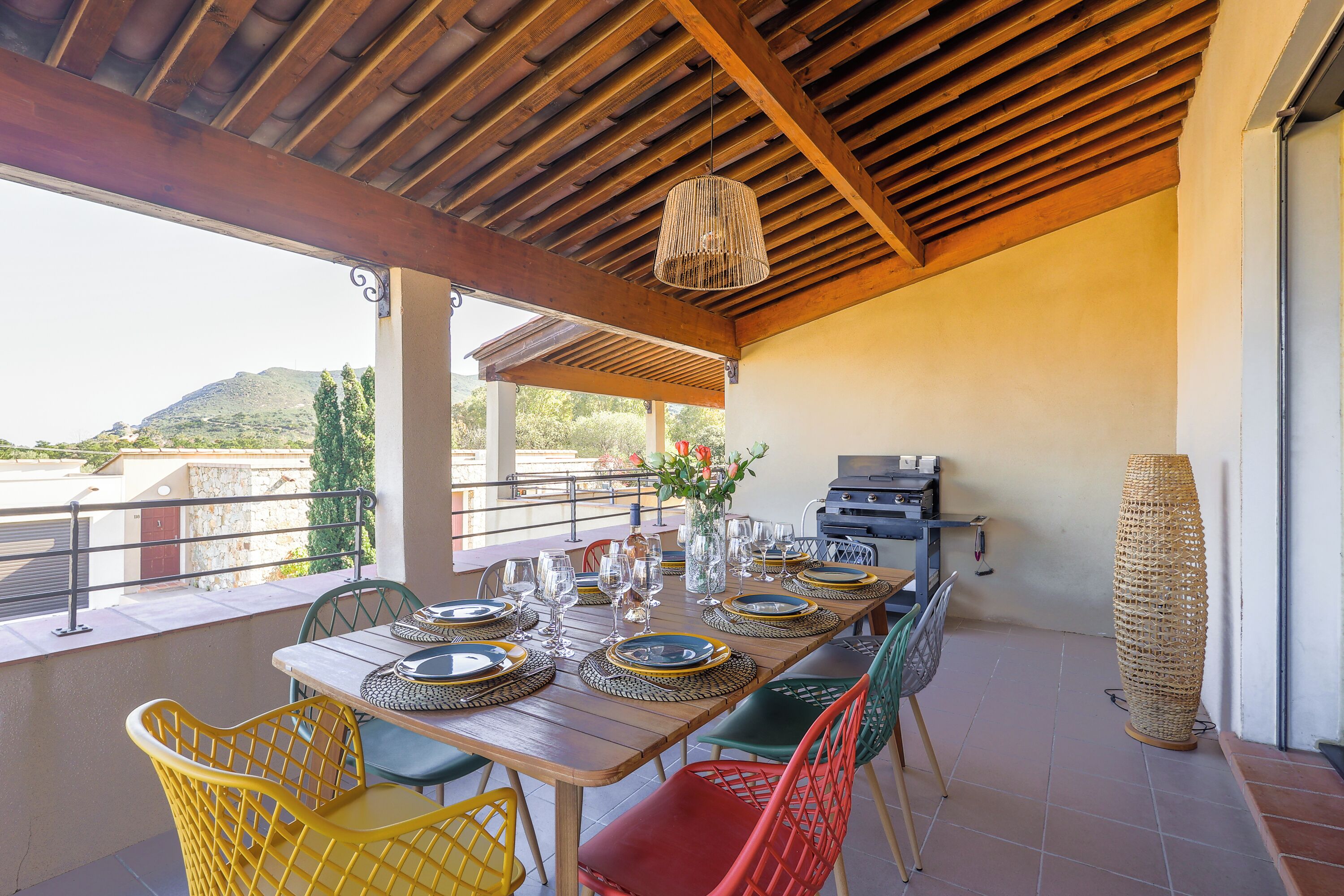 A covered outdoor dining area with a wooden table set for six, surrounded by colorful chairs.