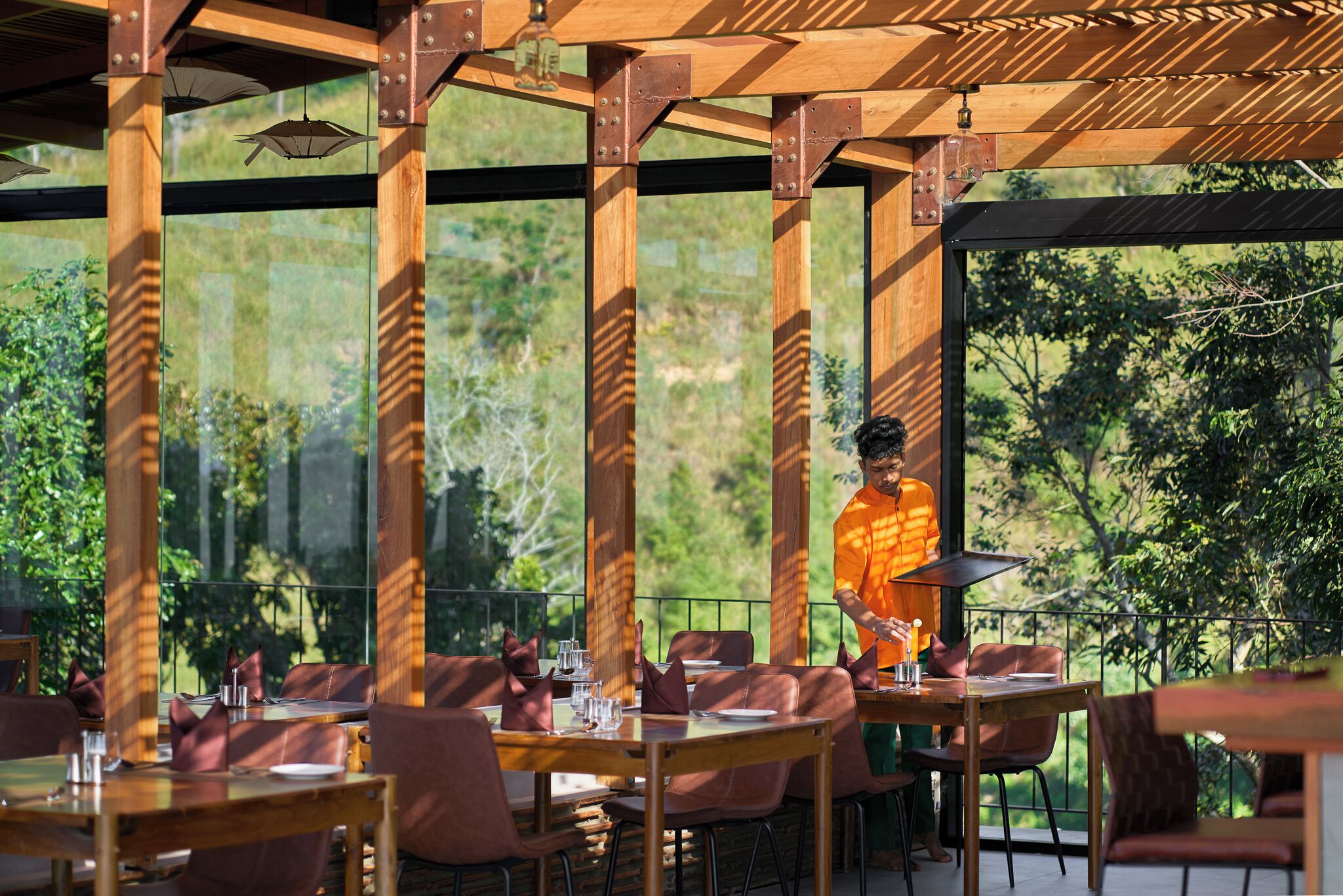 An outdoor dining area with wooden tables and chairs, large windows offering a view of greenery, and a person in an orange shirt working near the tables.