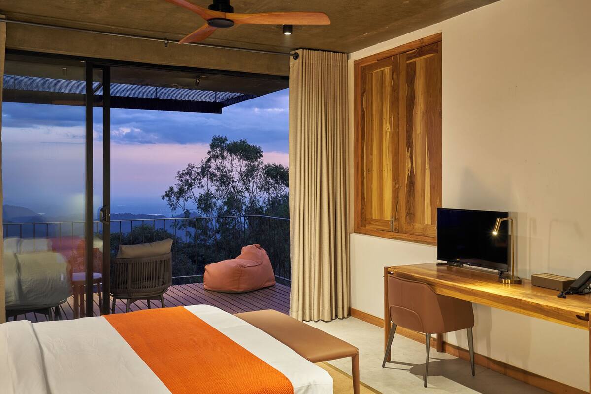 A bedroom with a bed, a desk, a television, and a balcony with a view of trees and mountains.