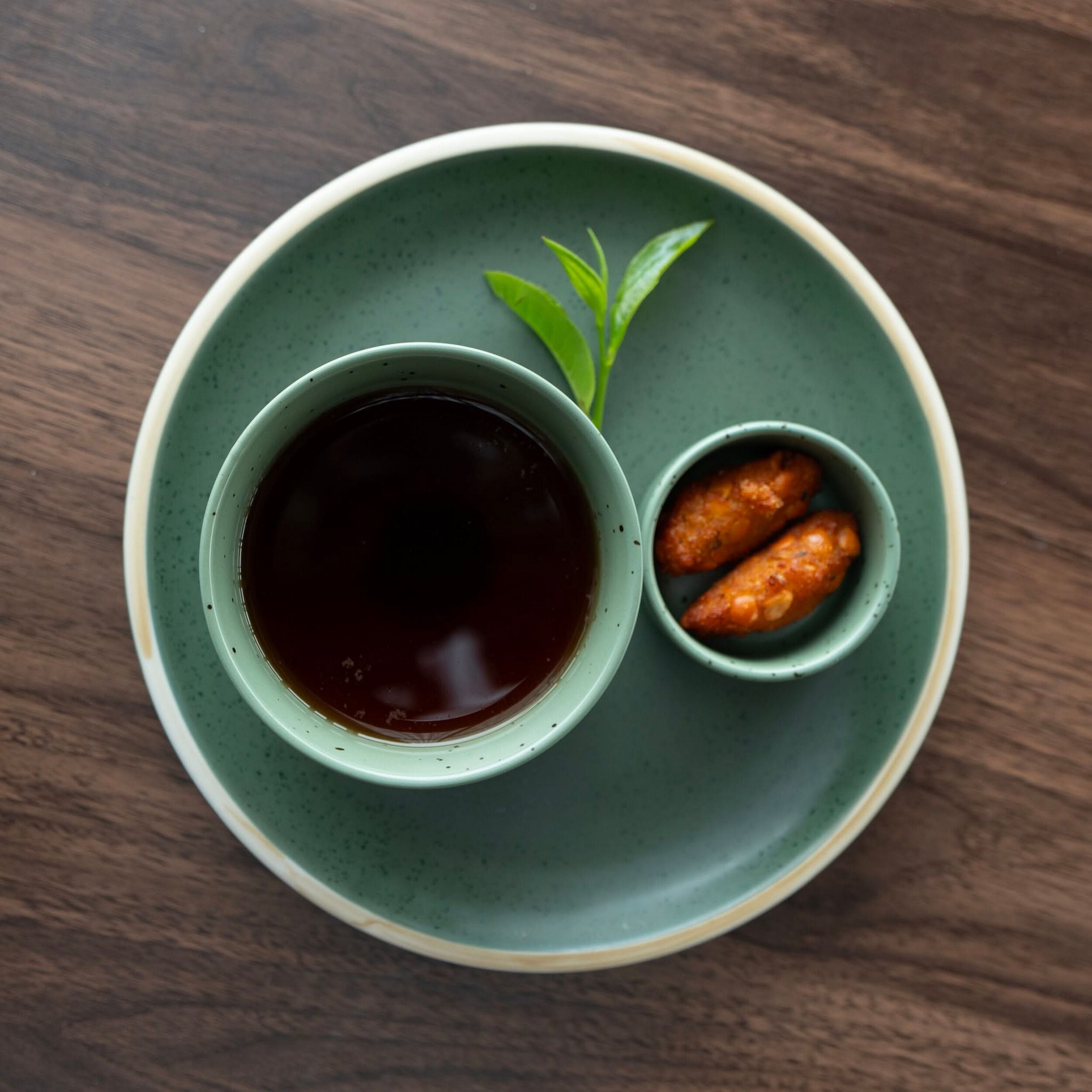 A green ceramic cup with a dark liquid, a small bowl with two orange items, and a green leaf on a round plate on a wooden surface.