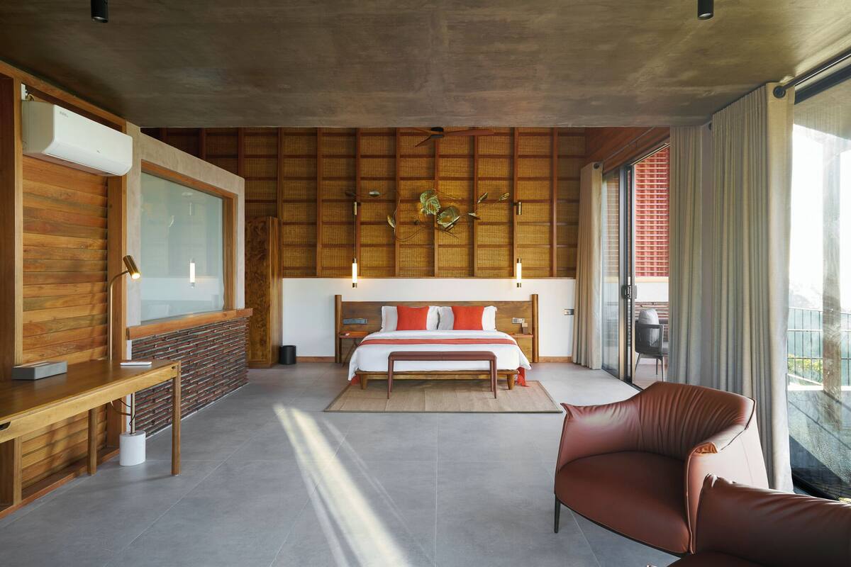 A modern bedroom with a wooden headboard, a large bed with red and white bedding, a bench, a desk, and a large window with curtains.