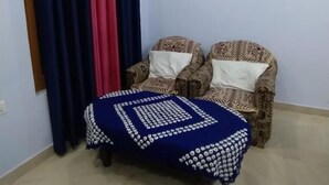 A room with a patterned armchair, a blue ottoman, and a curtain rod with multiple curtains. - The Gangotri Homestay By Preet rS (Rajgarhi)