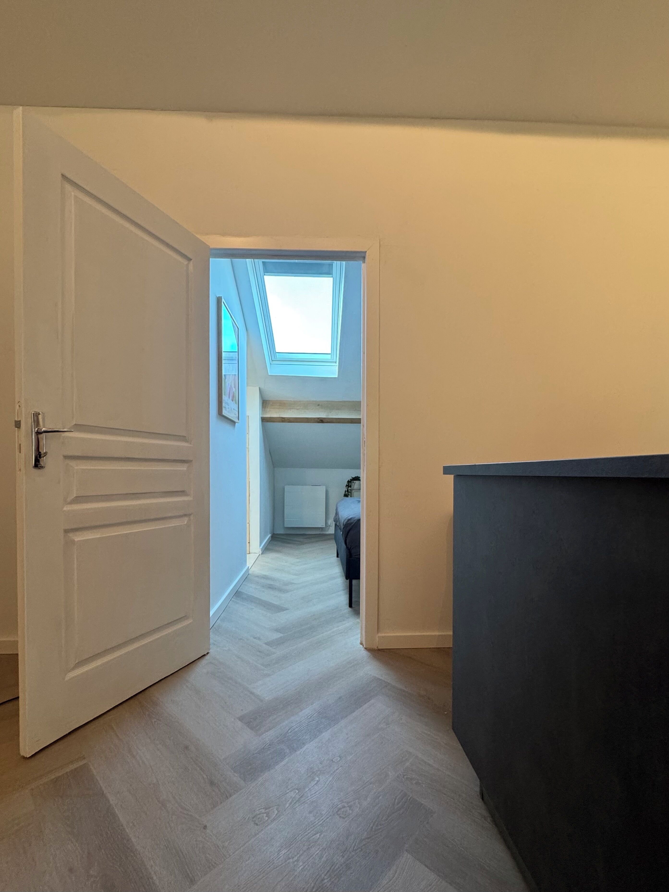 Een gang die leidt naar een slaapkamer met dakraam, radiator en bed.