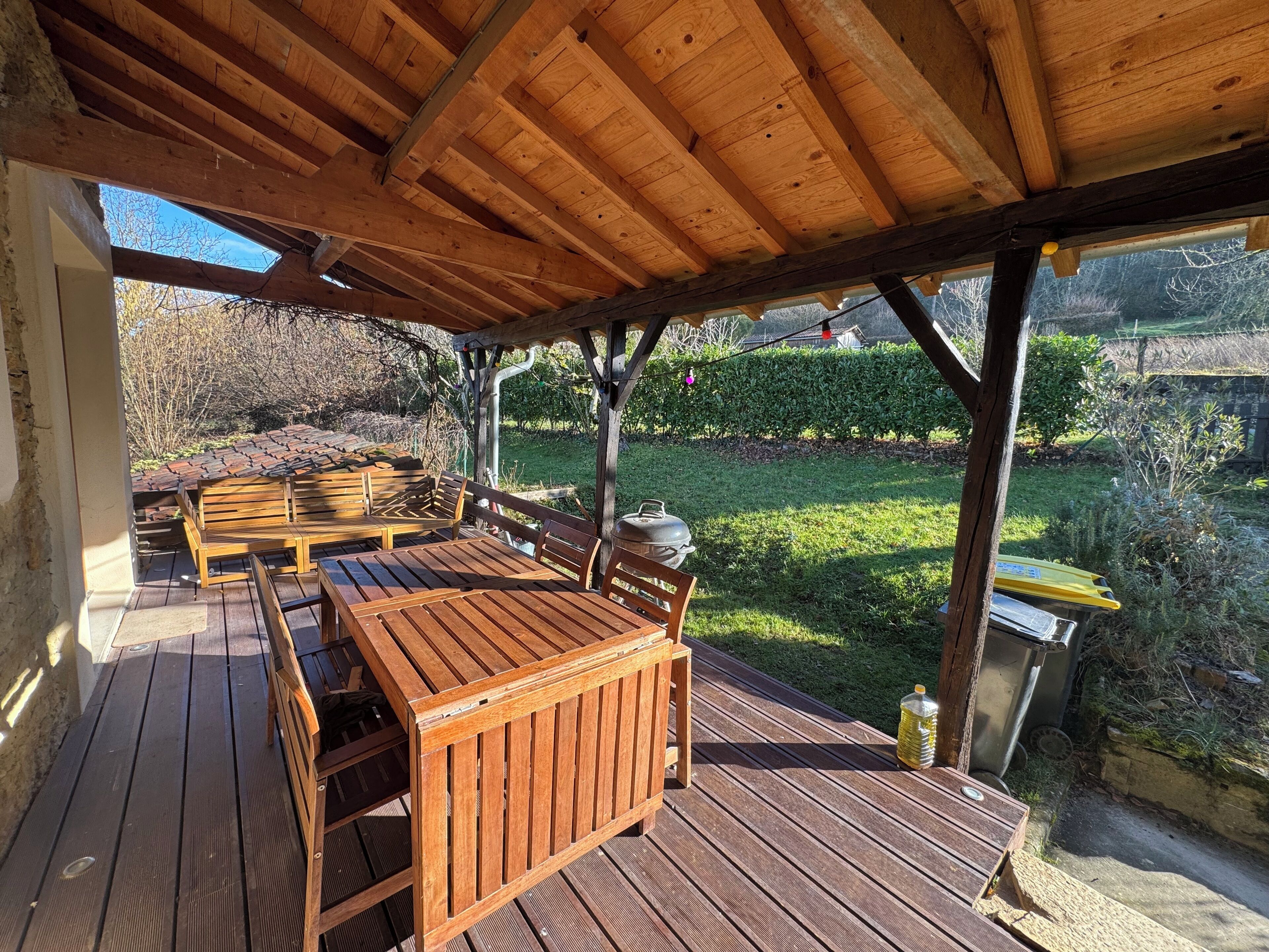 Una terraza de madera con una zona de estar cubierta, una parrilla y vistas a un jardín.