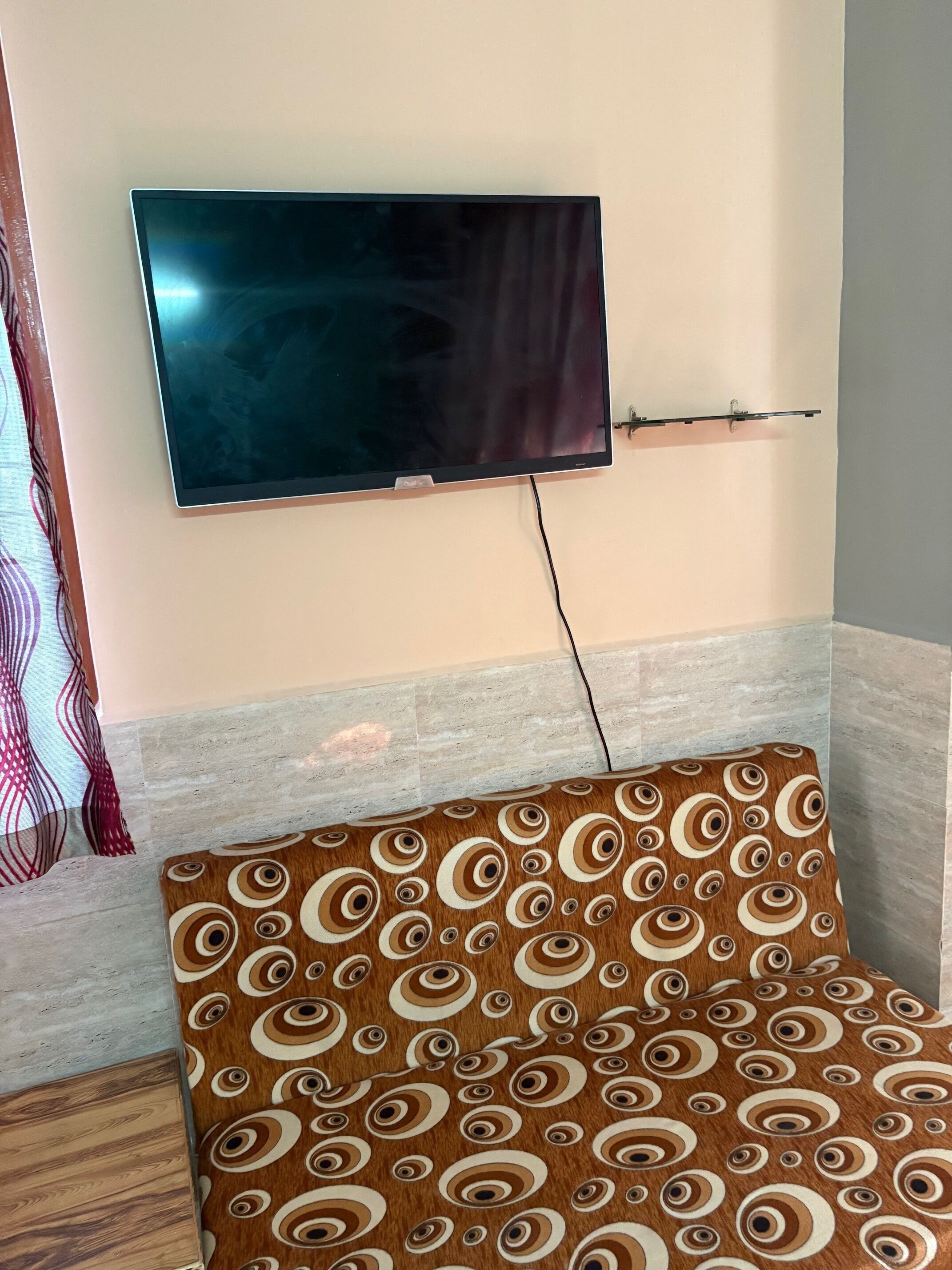 Una habitación con un televisor de pantalla plana montado en la pared, un cojín estampado en un sofá y un suelo de baldosas.