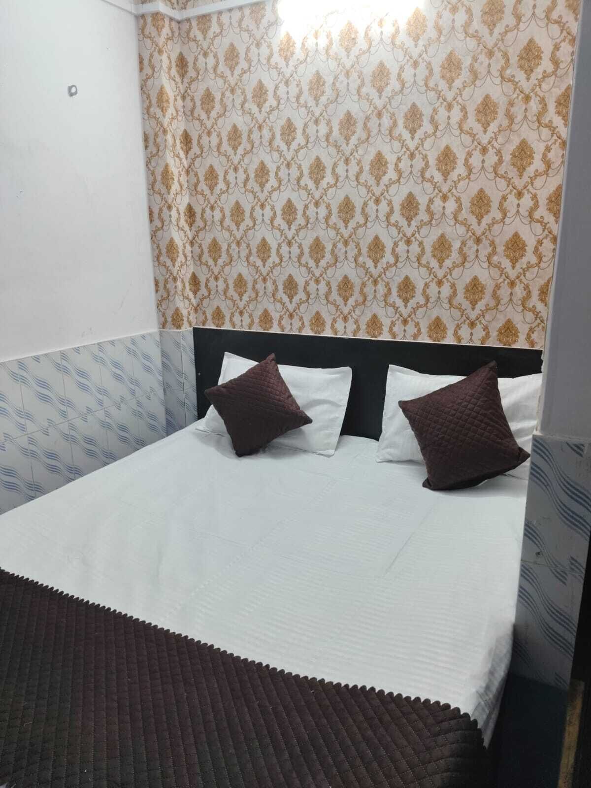 Una cama individual con colchón blanco y dos almohadas marrones, cabecera negra y papel tapiz estampado con diseño dorado y blanco.