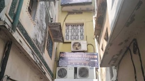 Une ruelle étroite bordée de bâtiments des deux côtés, avec une enseigne indiquant « Riverview Haven Guest House & Café », et plusieurs unités de climatisation fixées au bâtiment.