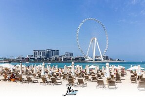 Una spiaggia con numerosi lettini e ombrelloni, una grande ruota panoramica e moderni edifici sullo sfondo.